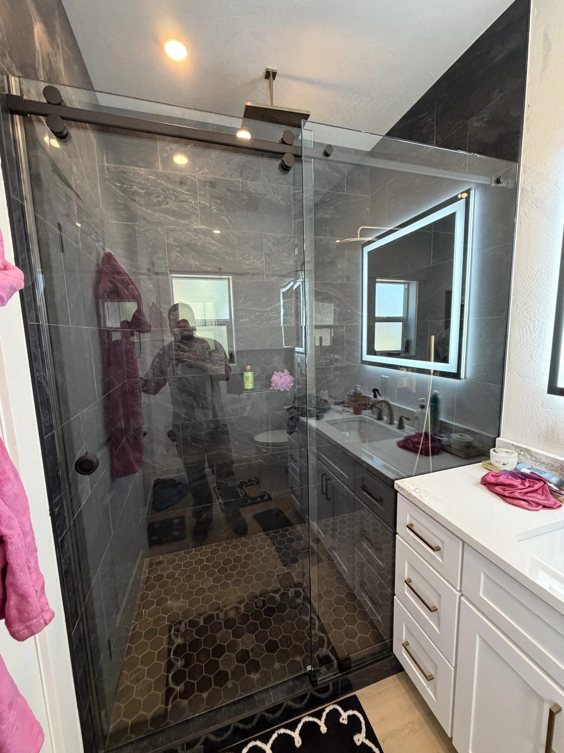 Frameless sliding shower door with black hardware beside a lit mirror